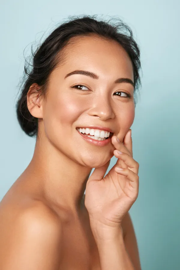A beautiful asian woman smiling against a light blue background, happy with her facial hair removal treatment in Miami.