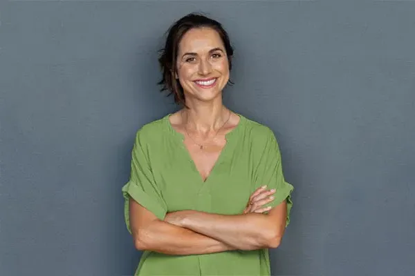 A woman in a green shirt stands smiling against a gray-blue wall, pleased with her hormone therapy from Vida Integrative Health of Miami in Miami.