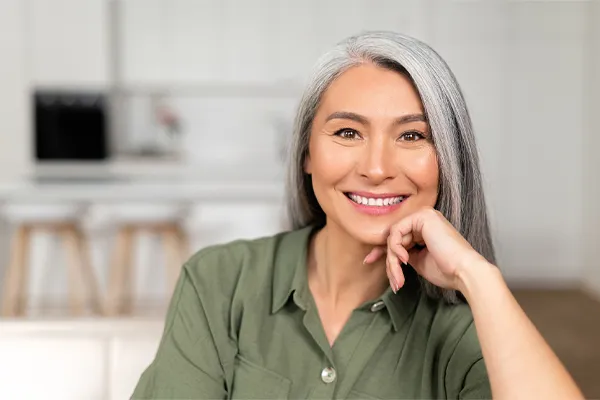 An asian woman with long gray hair and a green shirt sitting and smiling on her couch, happy with her menopause care from [NAME} in Miami.