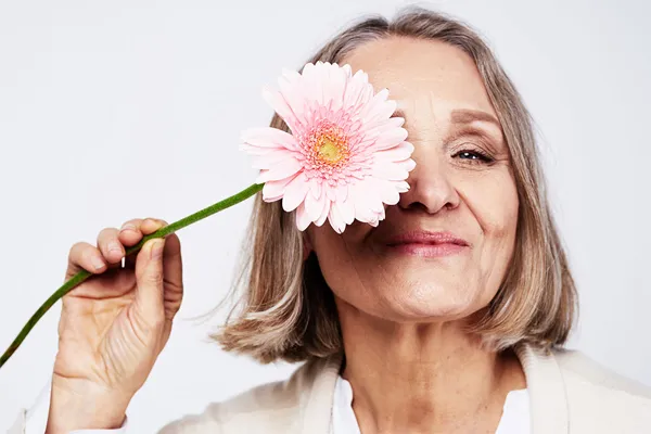 A smiling older woman holds a pink flower in front of her eye playfully, happy to have received menstrual dysfunction care from Vida Integrative Health of Miami in Miami.