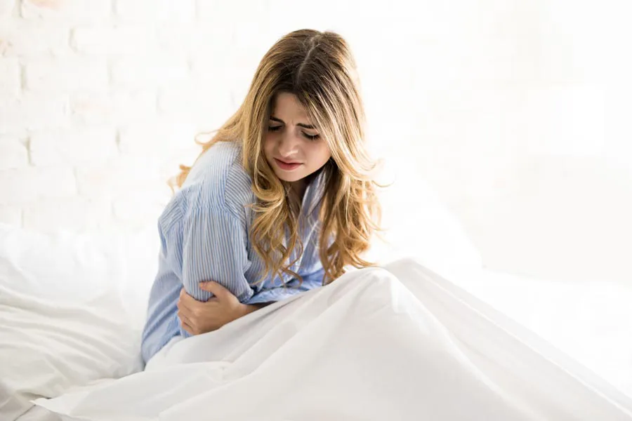 A woman with long blonde hair sits in her bed holding her stomach in pain, considering getting treatment for PMS from Vida Integrative Health of Miami in Miami.