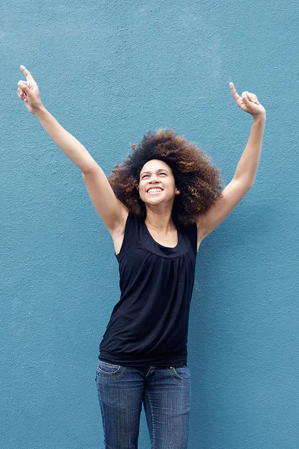 A woman in a dark blue tank top standing in front of a bright blue wall in Miami, raising her arms in celebration of relief from PMS.