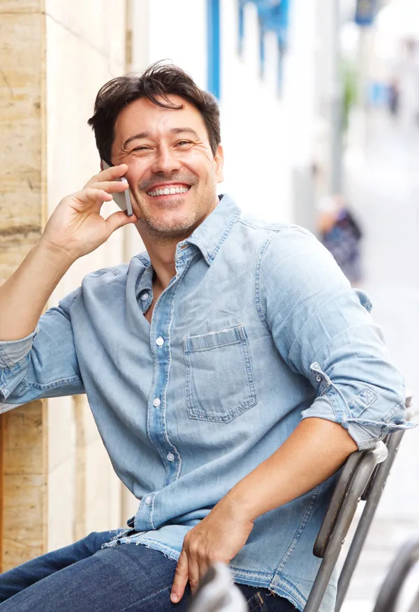 A man in a light blue shirt sits at a cafe laughing and talking on the phone, happy to have received peptides from Vida Integrative Health of Miami in Miami.