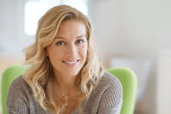 A woman with long blonde hair and a gray shirt sitting in a green chair and smiling, happy with her perimenopause treatment from Vida Integrative Health of Miami in Miami.