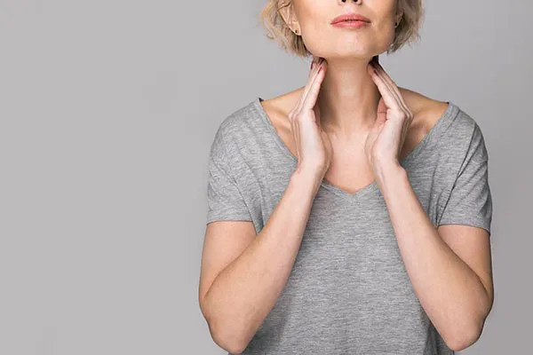 A woman in a gray t-shirt standing against a gray background massage the top of her neck, considering getting care for thyroid imbalance from Vida Integrative Health of Miami in Miami.