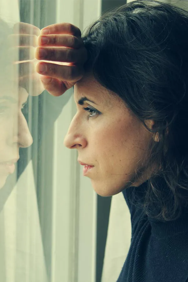 A brunette woman in a thin dark turtleneck looks out the window in Miami with a serious expression, suffering from PCOS.