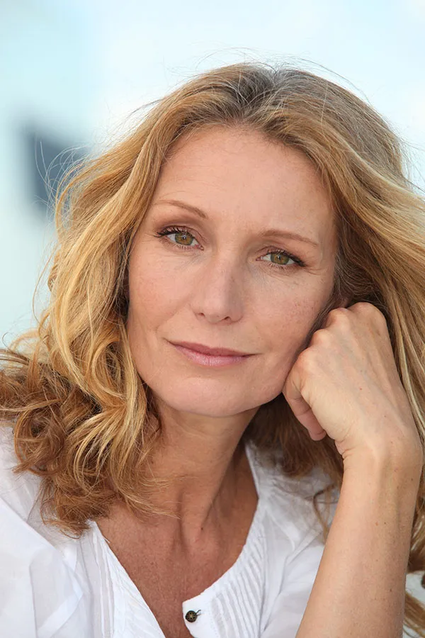 A beautiful blonde woman in a white blouse resting her head on her hand, living with Perimenopause in Miami.